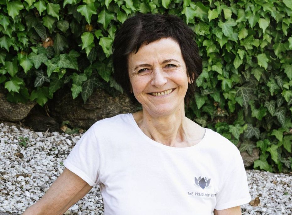 Portrait einer Mitarbeiterin im Preidlhof Eine lächelnde Mitarbeiterin steht vor einer mit Efeu bewachsenen Steinmauer und trägt weiße Kleidung mit Logo.