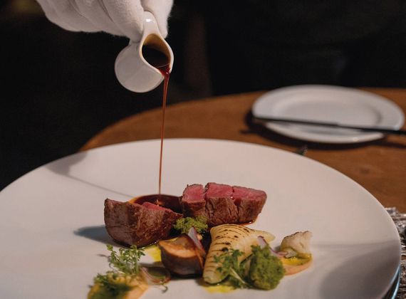 A gloved hand pours sauce over a plated gourmet beef dish with vegetables on a white plate.