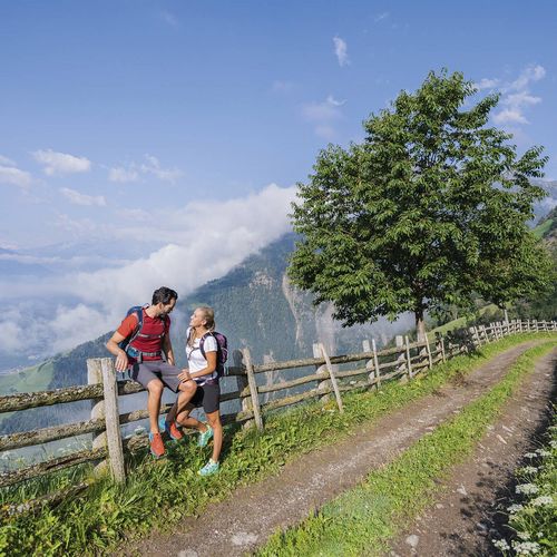 Zwei Wanderer stehen an einem Holzgeländer auf einem Bergweg mit Blick ins Tal und auf die umliegenden Berge.