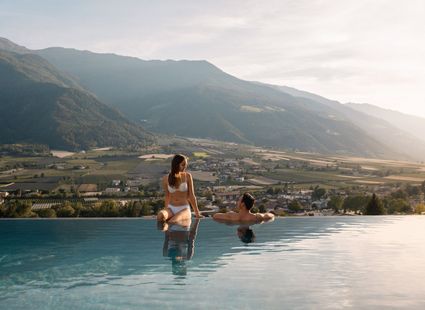 Zwei Frauen sitzen am Rand eines Infinity-Pools mit Blick über das Tal und die Berge.