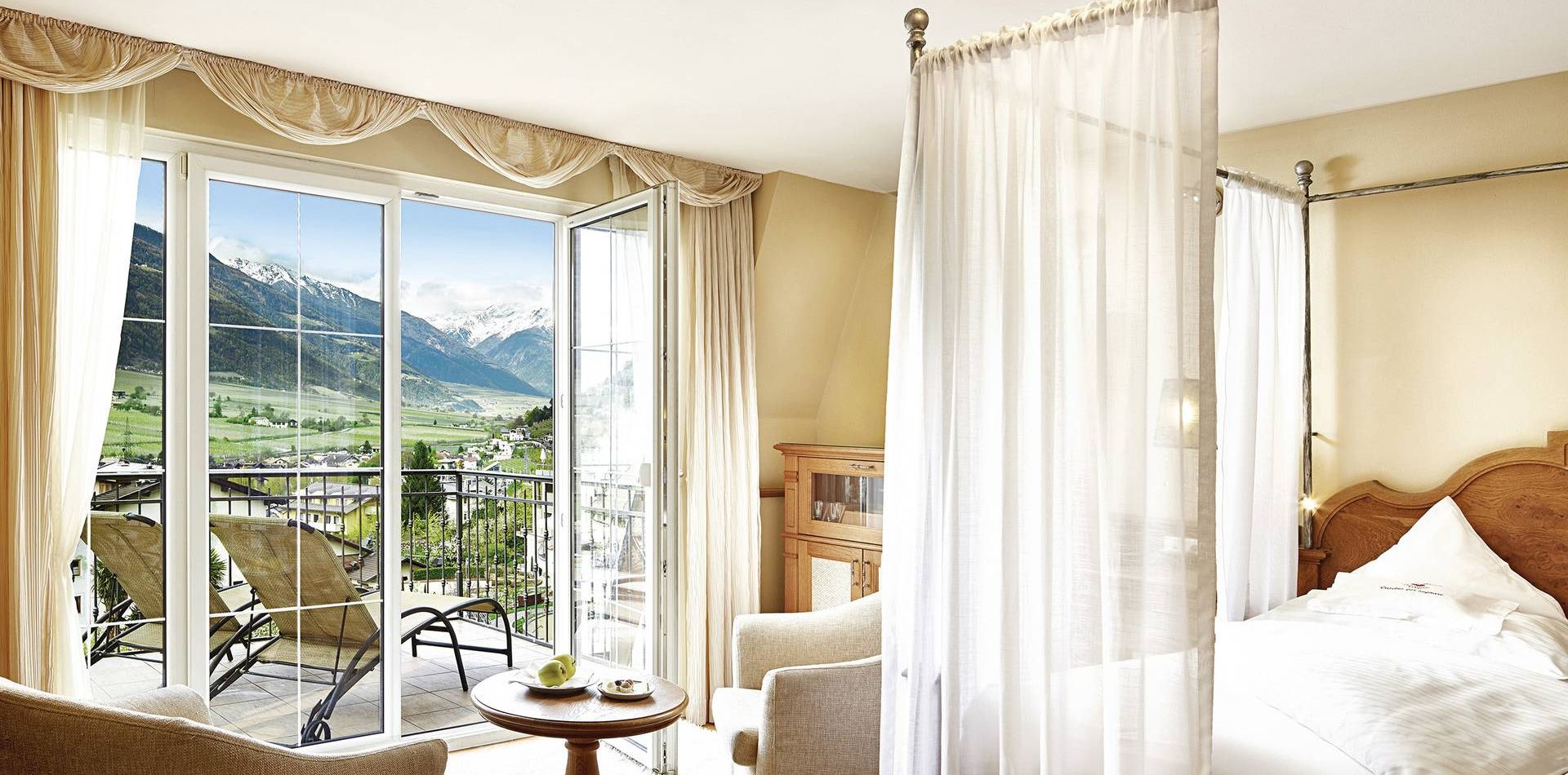 Una luminosa suite di lusso con letto a baldacchino, tenda leggera, poltrona e porte aperte sul balcone verso le montagne.
