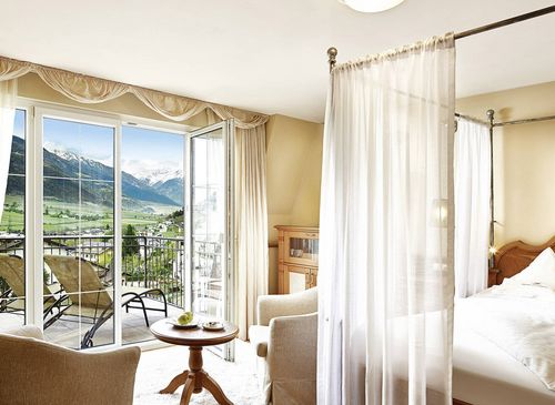 Luxury suite with balcony and mountain view A bright luxury suite with a four-poster bed, sheer canopy curtain, armchair, and open balcony doors facing the mountains.