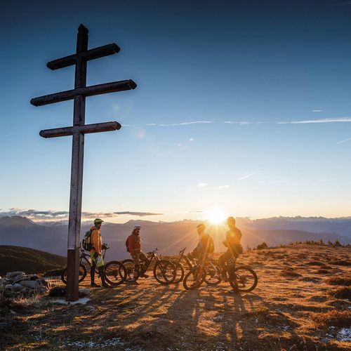 Mehrere Mountainbiker stehen auf einem Berggipfel neben einem Wegweiser bei Sonnenuntergang.