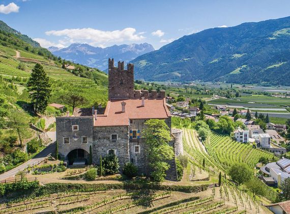 Ein mittelalterliches Schloss steht über terrassierten Weinbergen in einer weiten Berglandschaft.