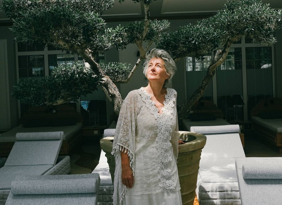 A woman in a white lace dress stands among sun loungers and trees in a shaded garden courtyard.