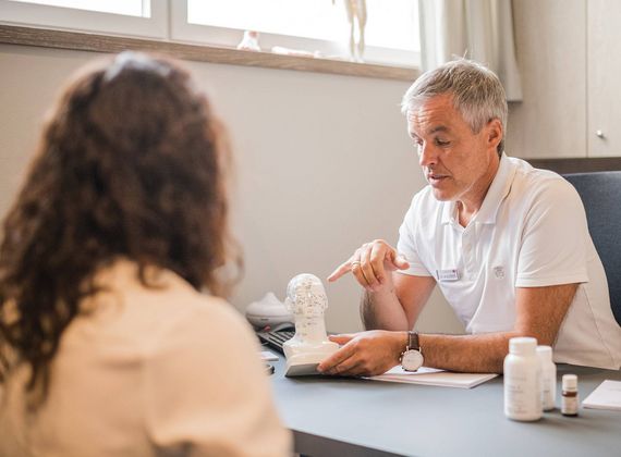 Ein Therapeut berät eine sitzende Person am Tisch im Medical Spa und zeigt ein Gerät auf einer Unterlage.