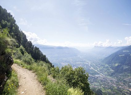 Ein schmaler Wanderweg führt am Hang entlang mit weitem Blick über das Tal und die Berge im Hintergrund.