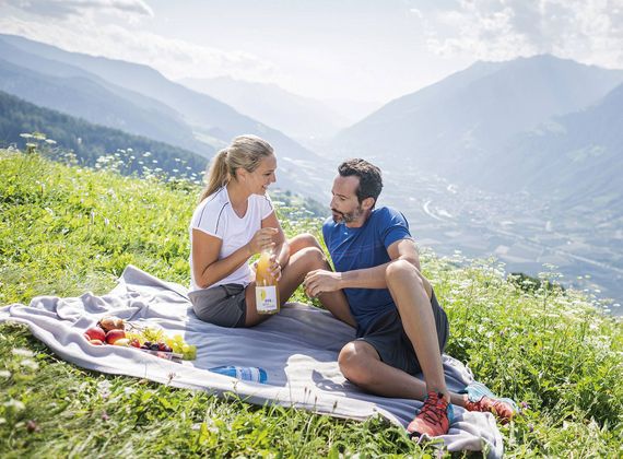 Ein Paar sitzt auf einer Decke auf einer Almwiese und macht ein Picknick mit Brot und Getränk vor Bergkulisse.