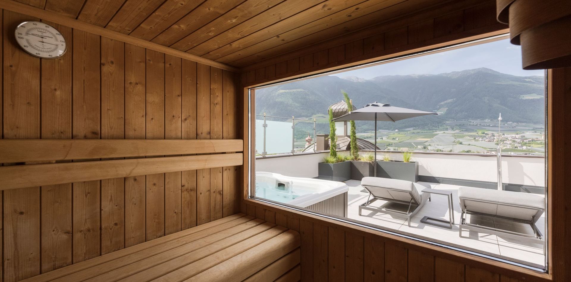Eine holzverkleidete Sauna mit großem Fenster zur Dachterrasse, Liegen und Blick auf die Berge.
