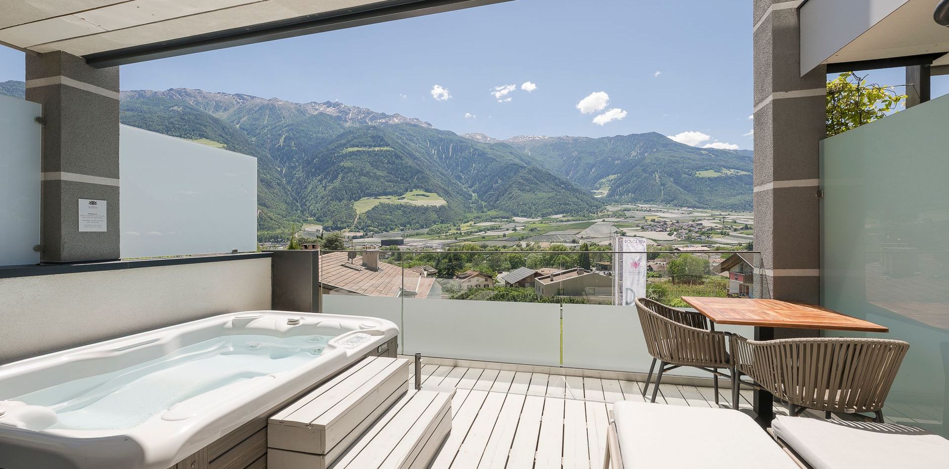 Eine moderne Suite-Terrasse mit Whirlpool, Liege und Tisch blickt auf die Berge und das Tal.