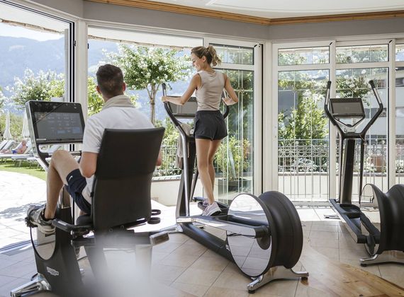 A man sits on a cardio machine while a woman uses a treadmill in a bright fitness room with mountain views.