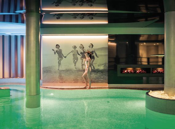 Ein Innenpool im Spa mit grün schimmerndem Wasser, Wandbild und Kamin in moderner Wellness-Atmosphäre.