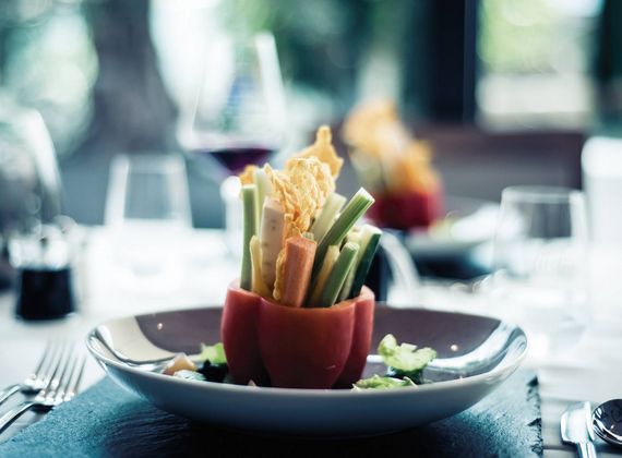 A plated vegetable appetizer sits on a dark table setting with blurred wine glasses in the background.