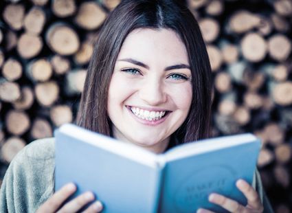 Eine lächelnde Frau hält ein blaues Buch vor einem Hintergrund aus gestapeltem Kaminholz.