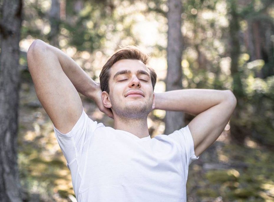 Ein Mann in weißem T-Shirt steht im Wald mit zurückgelehnten Armen und geschlossenen Augen.