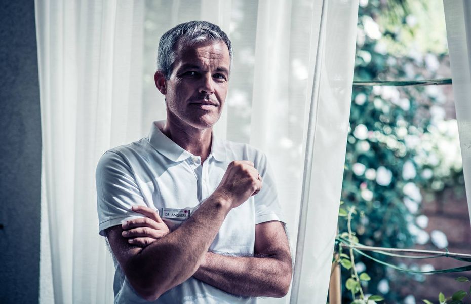 Ein Mitarbeiter im weißen Poloshirt steht vor einem Fenster mit Gardinen und blickt in die Kamera.