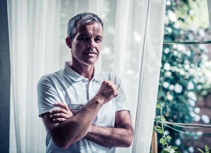 Ein Mitarbeiter im weißen Poloshirt steht vor einem Fenster mit Gardinen und blickt in die Kamera.