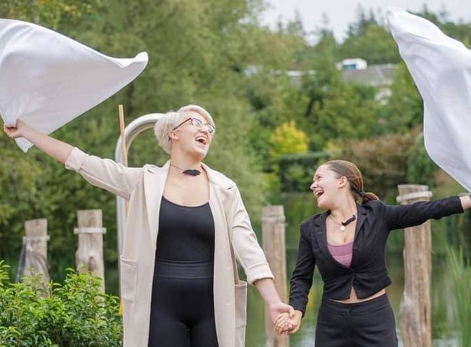 Zwei Frauen lachen und schwenken im Garten weiße Tücher, während sie sich an den Händen halten.