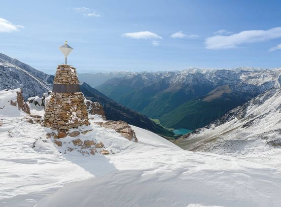 Ein Steinmarterl steht auf einem verschneiten Gipfel mit Blick auf das Tal und die umliegenden Berge.