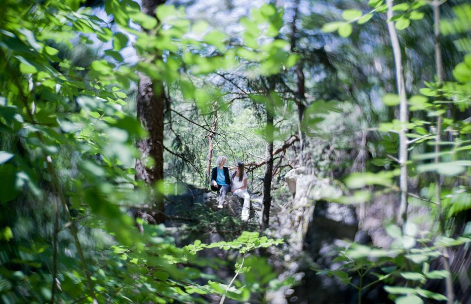 Wandern im Grünen beim Preidlhof in Naturns Zwei Personen sitzen auf einem Felsen im Wald, umgeben von grünem Laub und Bäumen.