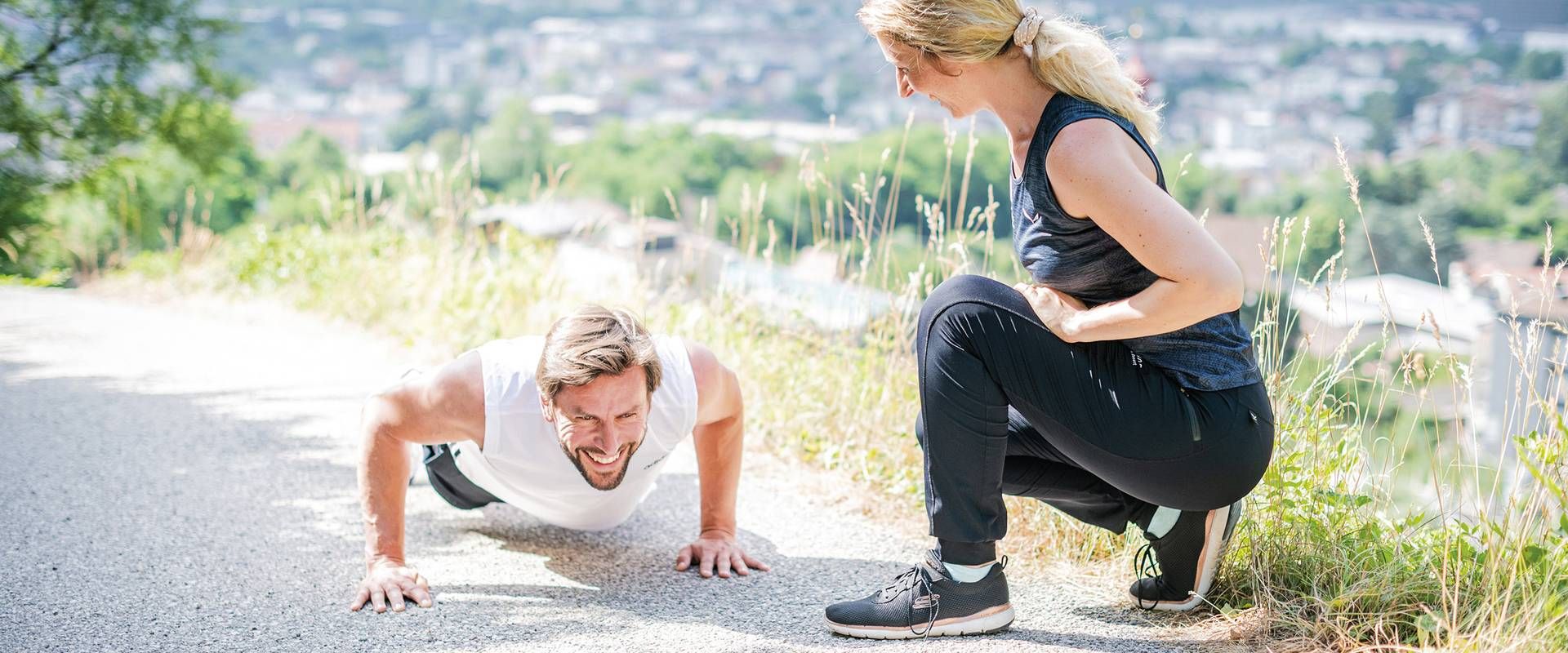 Ein Trainer macht Liegestütze neben einer sitzenden Frau auf einem Weg im Freien, mit Blick auf die Stadt im Hintergrund.