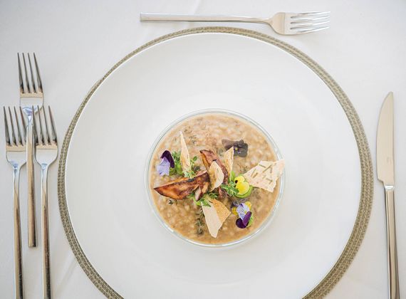 Un risotto gourmet impiattato con erbe, funghi e scaglie di formaggio su una mise en place bianca.