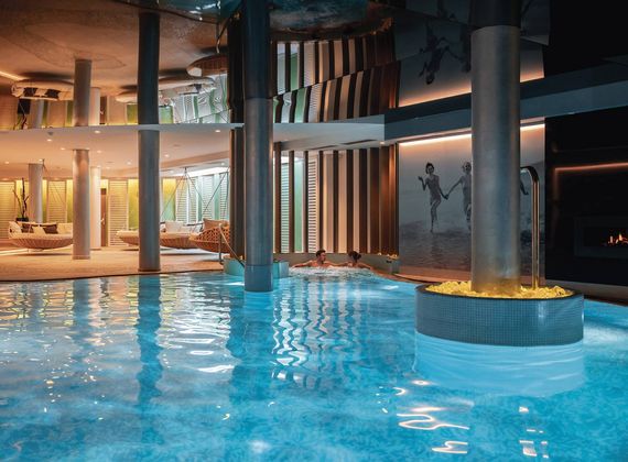 Ein Innenpool mit warm beleuchteten Liegen und einer flammenden Feuerstelle im Wellnessbereich.