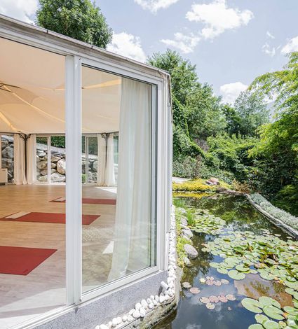 Ein heller Yoga-Pavillon mit Holzboden liegt am Rand eines Teichs mit Seerosen und Garten.