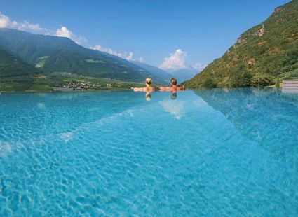 Zwei Gäste sitzen im Infinity-Pool und blicken auf das Bergpanorama über dem Tal.