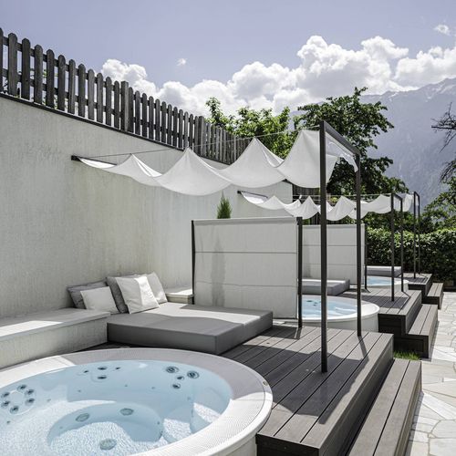 Ein runder Whirlpool steht auf einer sonnigen Terrasse mit Liegen, Sonnensegeln und Blick ins Grüne.