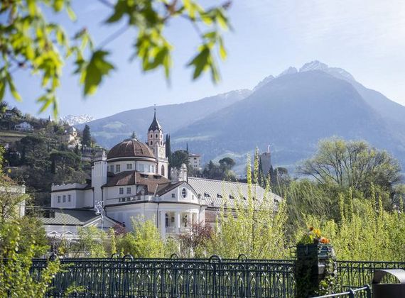 Eine Kirche mit Kuppel und Turm liegt vor einem Bergpanorama, eingerahmt von Bäumen und einem Zaun.