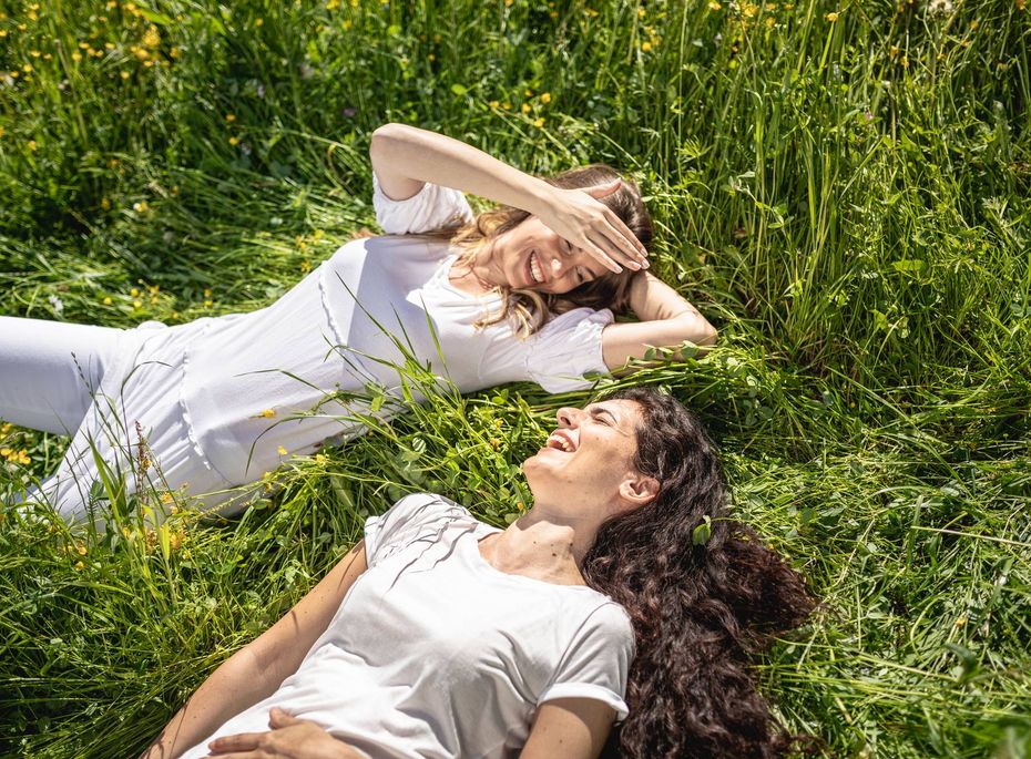 Zwei Frauen liegen im hohen Gras und lachen entspannt nebeneinander.