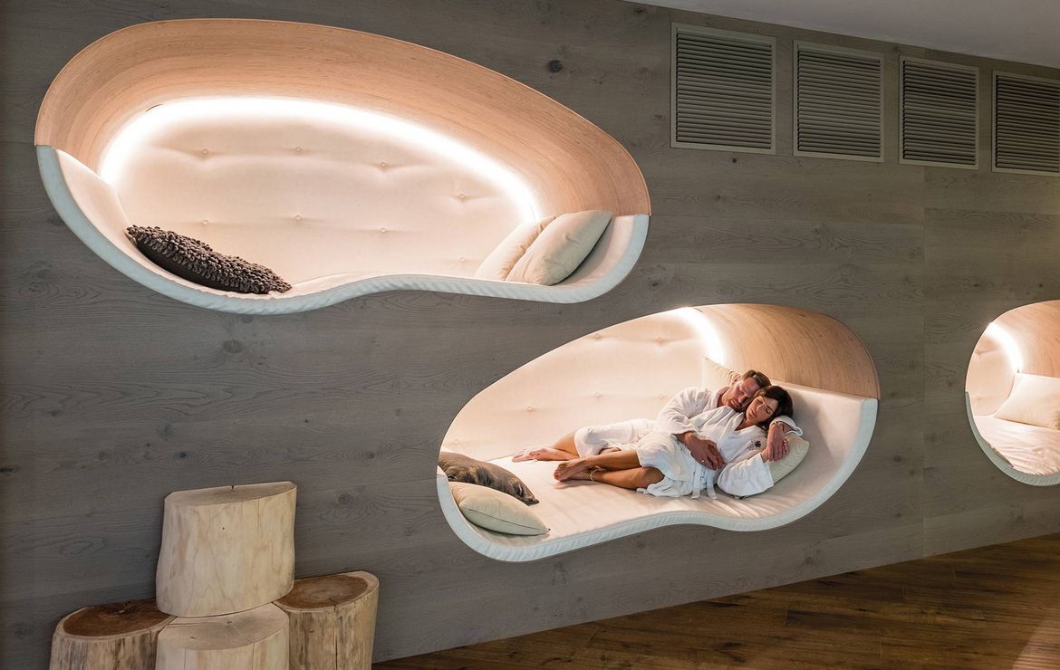 Two people lie together on a curved relaxation lounger in a spa room with soft lighting and wood flooring.