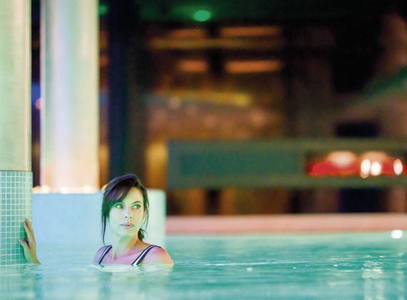 A woman stands in an indoor spa pool, holding the pool edge and looking to the side.