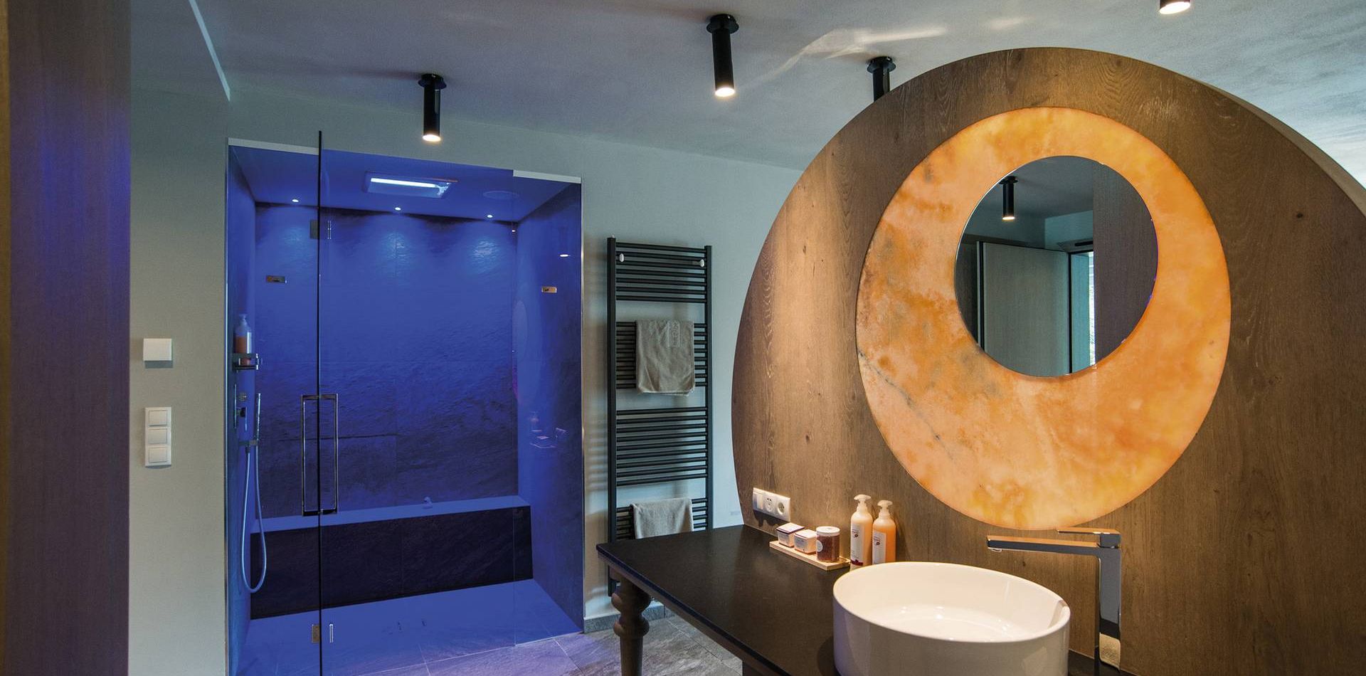 A suite bathroom with a round basin, dark vanity, and walk-in shower beside a blue-lit glass enclosure.