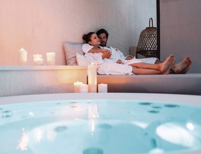Ein Paar sitzt gemeinsam auf einer Liege am Wellnessbecken, umgeben von Kerzen und ruhiger Spa-Atmosphäre.