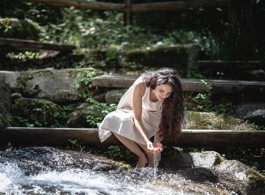Eine Frau beugt sich an einem Naturbach und lässt Wasser über ihre Hände laufen.