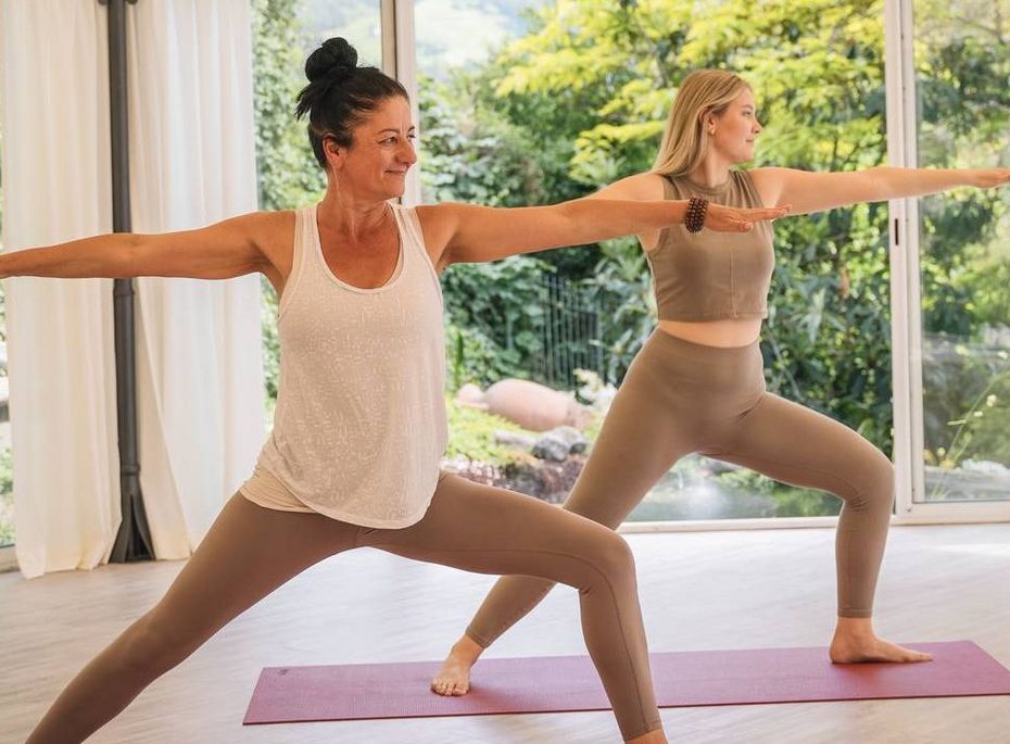 Yoga-Kurs im Preidlhof Wellnesshotel Zwei Frauen üben Yoga auf Matten in einem hellen Raum mit großen Fenstern und Blick ins Grüne.