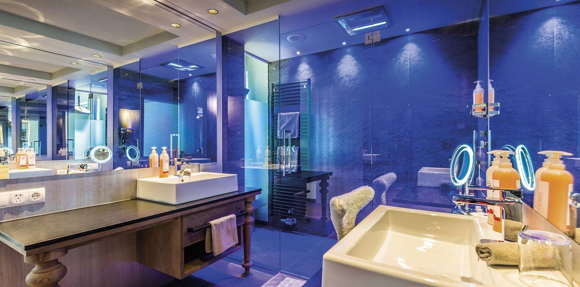 A bathroom with a double sink, large mirror, blue glass shower enclosure and dark wood vanity.