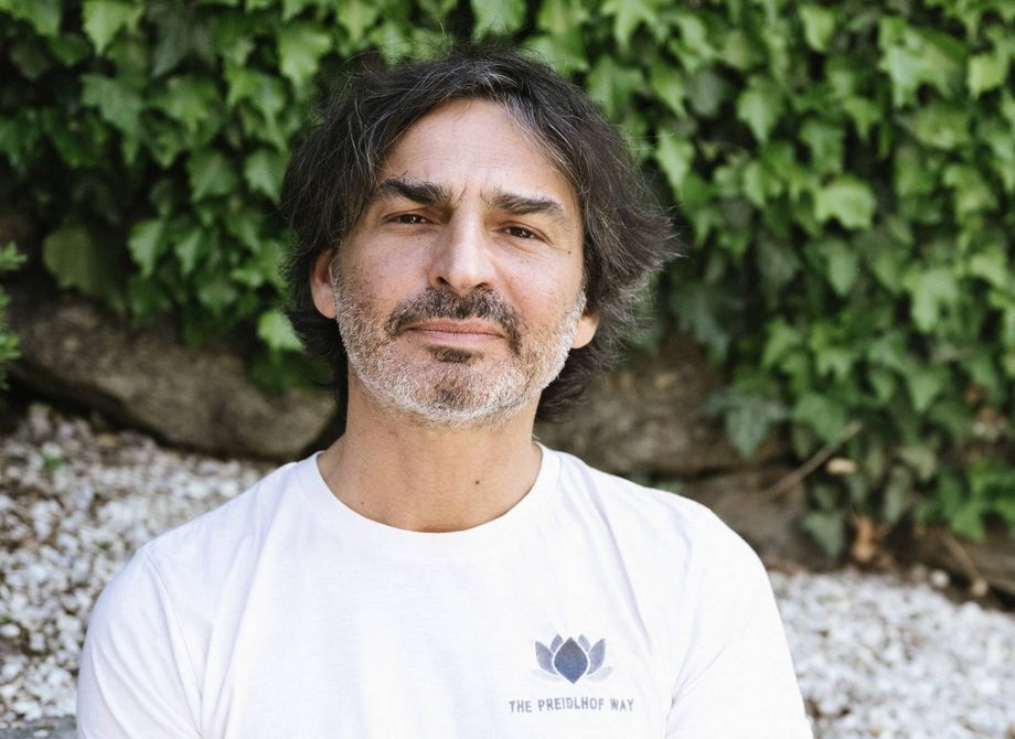 A wellness expert with dark hair and a beard stands in front of green ivy wearing a white Preidlhof Way T-shirt.