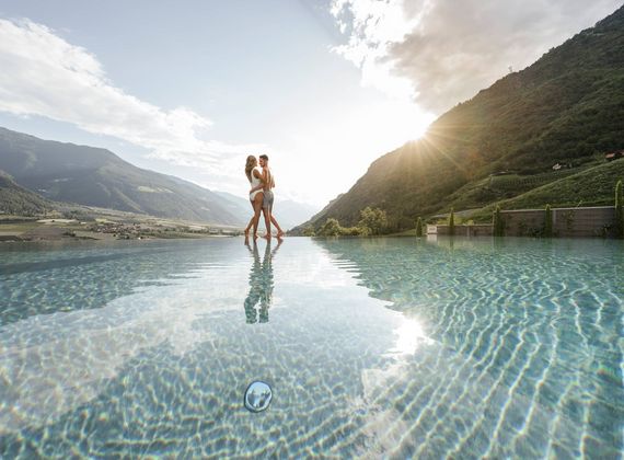 Zwei Frauen stehen im Infinity-Pool mit Blick auf die Berge und die tief stehende Sonne.