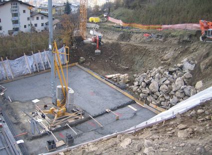 Eine Baustelle mit Betonfundament, Baumaschinen und aufgeschütteten Felsbrocken am Hang.
