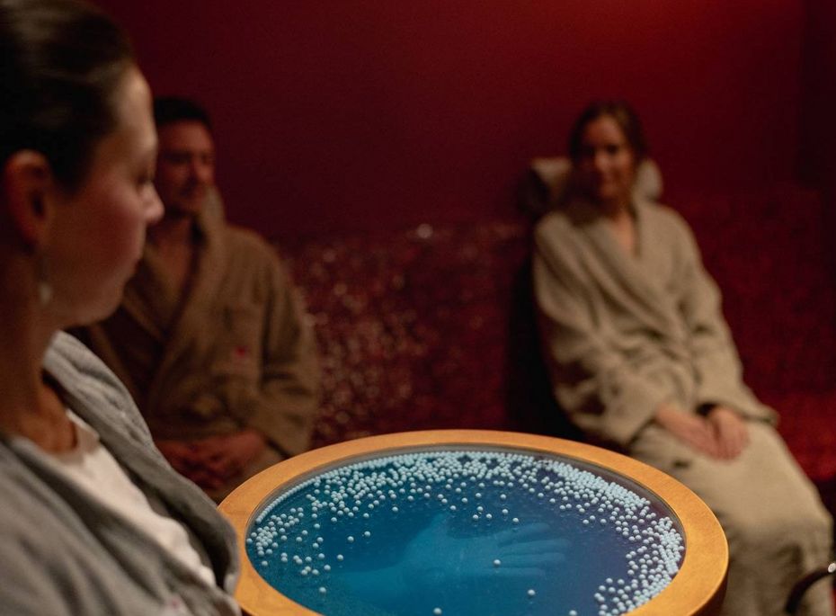 Eine Mitarbeiterin serviert einer sitzenden Frau und zwei weiteren Gästen ein Fußbad in einem warm beleuchteten Spa-Raum.