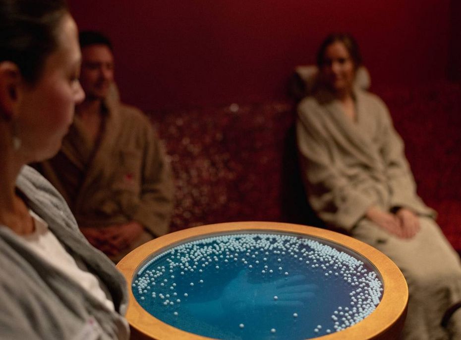 Spa-Anwendung mit Fußbad im Preidlhof Eine Mitarbeiterin serviert einer sitzenden Frau und zwei weiteren Gästen ein Fußbad in einem warm beleuchteten Spa-Raum.