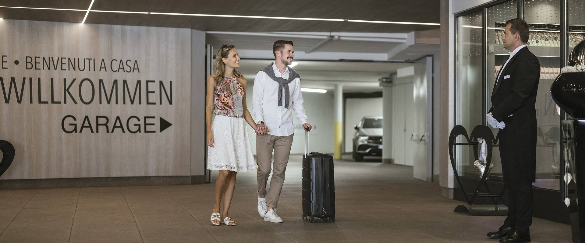 Ein Paar mit Koffer steht in der Hotelgarage und wird vom Concierge empfangen.