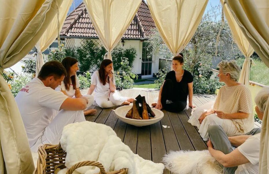 Wellness-Runde am Feuer im Preidlhof-Garten Fünf Personen sitzen in einer Wellness-Runde um eine Feuerschale im Garten unter einem hellen Pavillon.