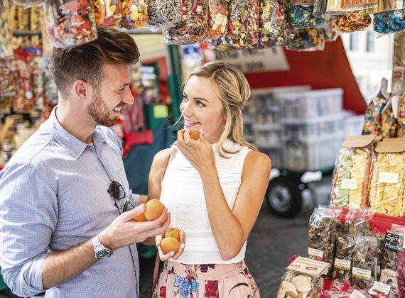 Ein Paar probiert Obst an einem Marktstand mit bunten Süßwaren und verpackten Snacks.