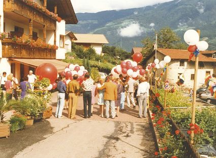 Eine Menschenmenge mit Luftballons steht auf einer Dorfstraße zwischen Häusern und Gärten vor Bergkulisse.