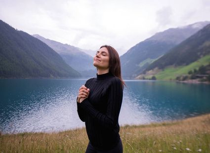 Eine Frau steht entspannt am Ufer eines Bergsees und genießt die ruhige Landschaft.