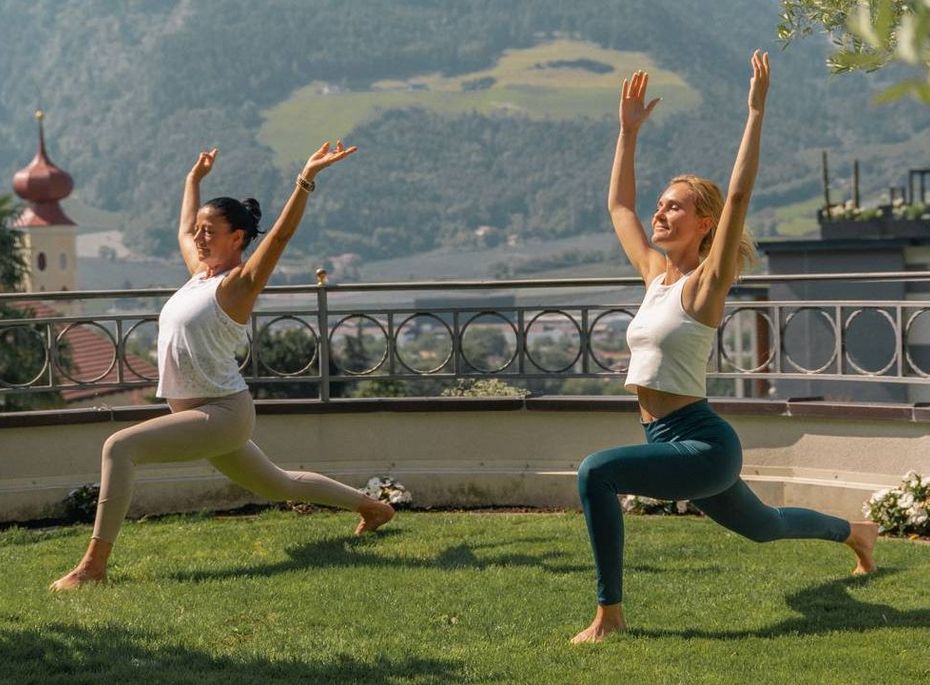 Zwei Frauen üben Yoga auf einer Terrasse mit Bergblick und Blick ins Tal.